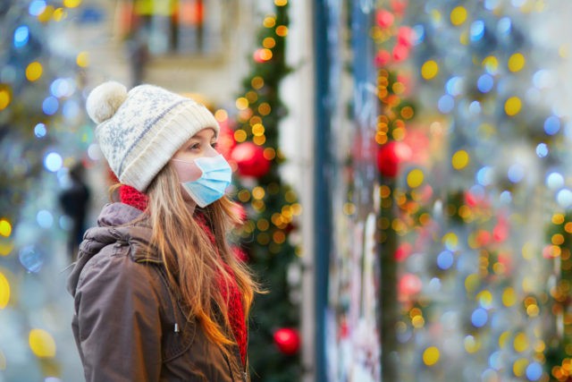 Girl wearing face mask on a Parisian street or at Christmas market looking at shop windows decorated for Christmas. Seasonal holidays during pandemic and coronavirus outbreak