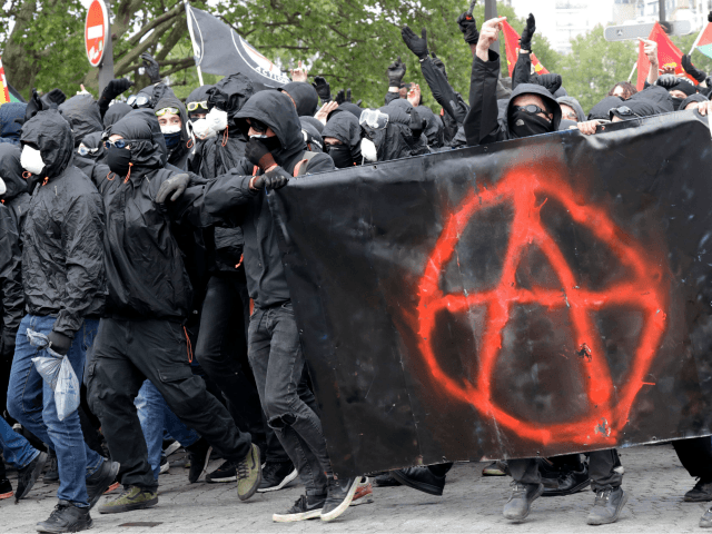 Protestors dressed in black and with the face covered, hold a banner picturing the symbol of the Anarchist as they take part in a march for the annual May Day workers' rally, in Paris, on May 1, 2018. (Photo by Thomas SAMSON / AFP) (Photo credit should read THOMAS SAMSON/AFP …