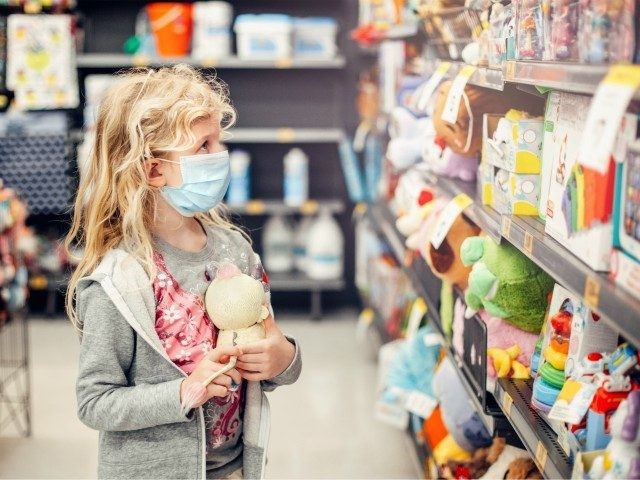 A new normal. Caucasian blonde girl in sanitary face mask shopping at toy store. Child wearing protective mask against coronavirus. Safety, health protection during covid-19 quarantine. - stock photo