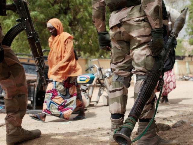 In this photo taken Wednesday, April 8, 2015, a woman walks past Nigerian Soldiers at a checkpoint in Gwoza, Nigeria, a town newly liberated from Boko Haram. Each day brings new reports of atrocities, with mass graves being discovered in towns seized back from the militants who had set up …