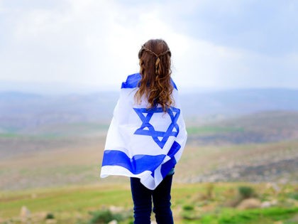 Little patriot jewish girl standing and enjoying great view on the sky, spring field and mountains with the flag of Israel wrapped around her. Memorial day-Yom Hazikaron and Yom Haâatzmaut concept.