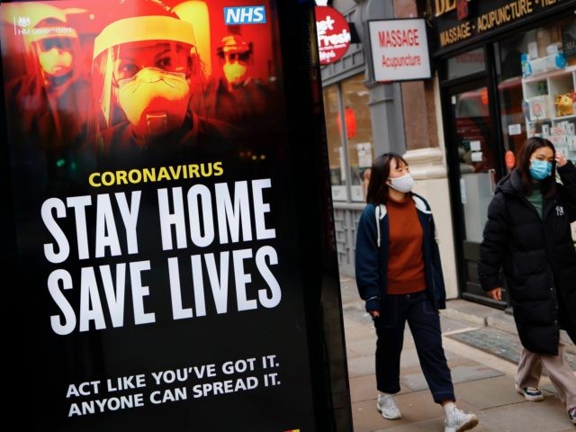Pedestrians wearing facemasks walk past NHS signage promoting "Stay Home, Save Lives" on a