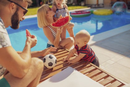 Young parents having fun spending hot sunny summer day outdoors with their children, eating slices of watermelon by the swimming pool