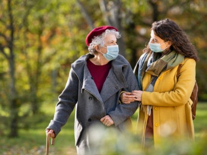 Lovely granddaughter walking with senior woman holding stick in park and wearing mask for safety against covid-19. Happy old grandmother enjoying walking in park with girl. Smiling elderly woman with happy caregiver in park relaxing after quarantine due to coronavirus outbreak and lockdown.