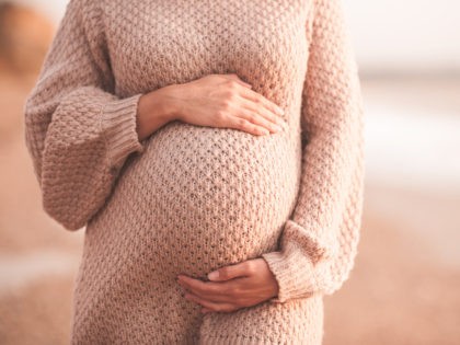 Pregnant woman wearing cozy knitted sweater holding tummy with hands outdoors close up. Mo