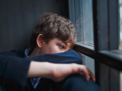 Pensive sad boy teenager in a blue shirt and jeans sitting at the window and closes his face with his hands. - stock photo