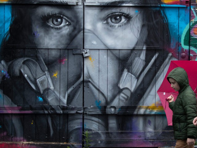 LONDON, ENGLAND - JUNE 11: A boy holding an umbrella walks past a mural of a woman wearing a mask in Shoreditch on June 11, 2020 in London, England. As the British government further relaxes Covid-19 lockdown measures in England, this week sees preparations being made to open non-essential stores …