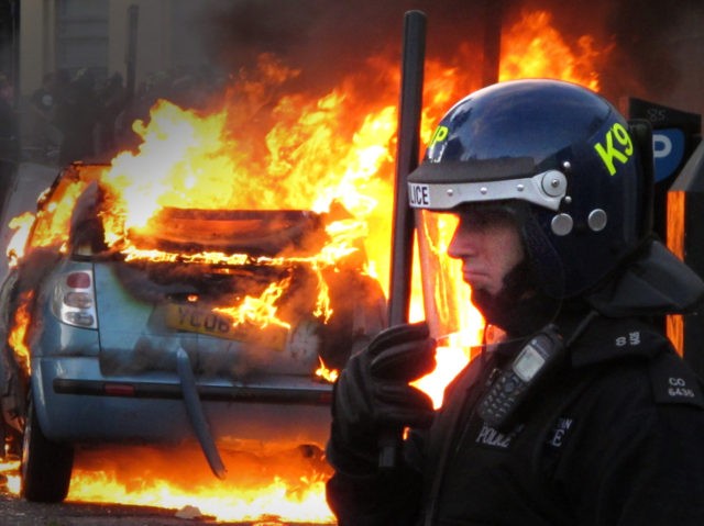 LONDON, ENGLAND - AUGUST 08: A police officer in riot gear stands near a burning car in Hackney on August 8, 2011 in London, England. Pockets of rioting and looting continues to take place in various boroughs of London this evening, as well as in Birmingham, prompted by the initial …