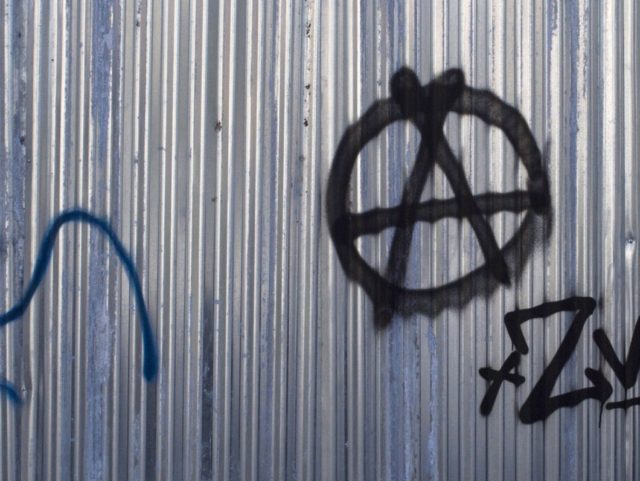 A man walks past a graffiti bearing the symbol of anarchism, an A for Anarchy in a circle, on March 17, 2017 in the Greek capital, Athens. Greek authorities on March 17, 2017 were trying to determine how a near-defunct militant group was able to sneak at least two parcel …