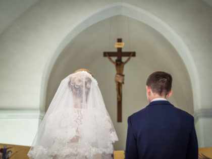 Photo taken during the wedding ceremony in Catholic church. Groom and bride stand before crucifix. Focus is on the crucifix. Newlyweds are in focus. Bride dressed in wedding dress and bridal veil. Groom dressed in suit and shirt. Photo taken from the back of bride and groom.