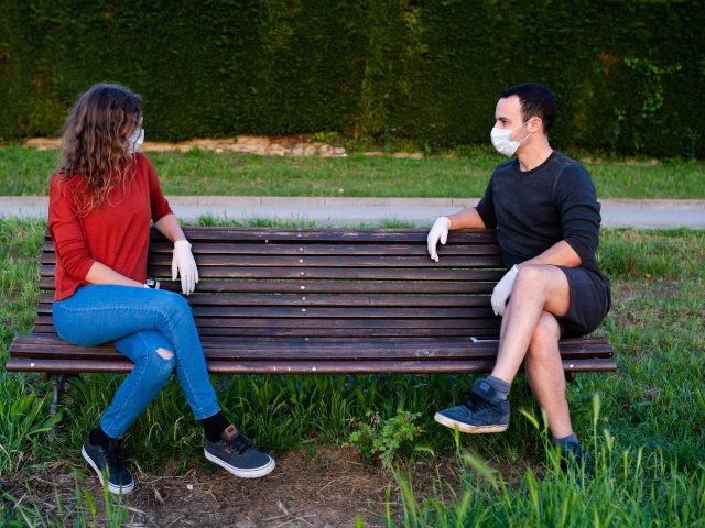 man and woman sitting on a bench in the park, keeping social distance wearing mask and gloves. Covid lifestyle - stock photo Covid 19 lifestyle coronavirus