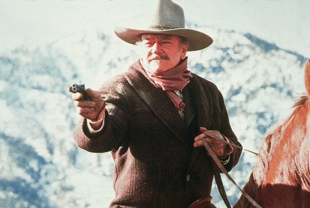 American actor John Wayne (1907 - 1979) aims a gun while riding horseback in a scene from