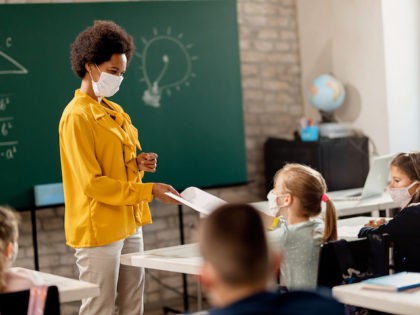 Happy black teacher and her students wearing protective face mask in the classroom. Teacher is giving them their test results.