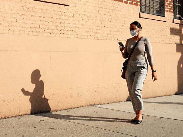 A woman wearing a protective mask walks while looking at a cellphone as the city continues