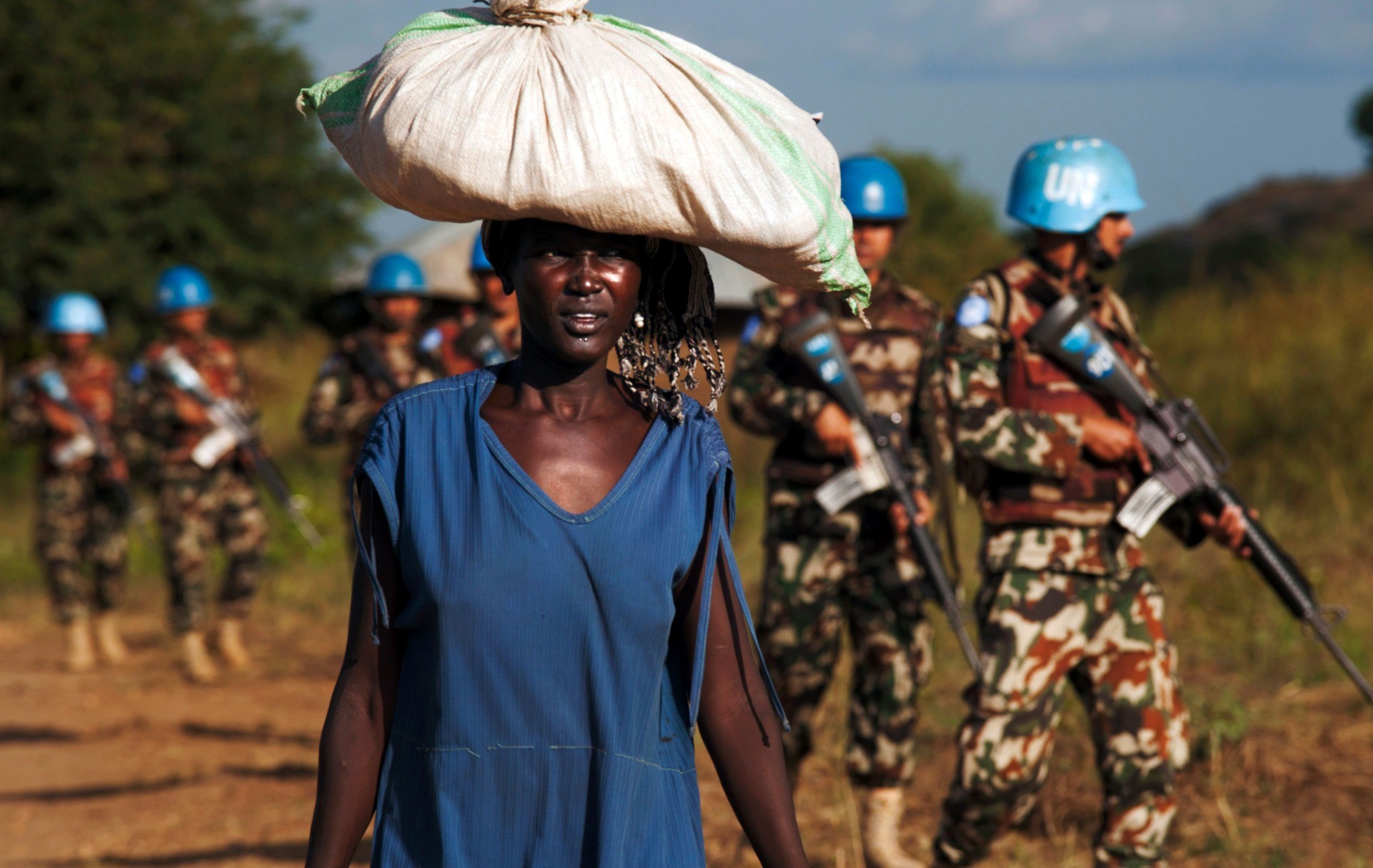 TOPSHOT - A displaced woman carries goods as United Nations Mission in South Sudan (UNMISS) peacekeepers patrol outside the premises of the UN Protection of Civilians (PoC) site in Juba on October 4, 2016. According to the UN, due to the increase of sexual violence outside the PoC, UNMISS has intensified its patrols in and around the protection sites, as well as in the wider Juba city area, sometimes arranging special escorts for women and young girls. / AFP / ALBERT GONZALEZ FARRAN (Photo credit should read ALBERT GONZALEZ FARRAN/AFP via Getty Images)