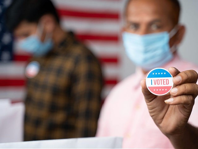 Man in medical mask showing I voted Sticker at polling booth with US flag as background