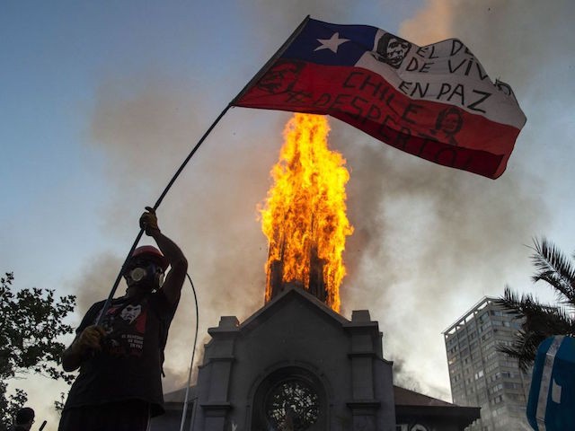 A demonstrator flutters a Chilean flag outside the burning church of Asuncion, set on fire by protesters, on the commemoration of the first anniversary of the social uprising in Chile, in Santiago, on October 18, 2020, as the country prepares for a landmark referendum. - Two churches were torched as tens of thousands of demonstrators gathered Sunday in a central Santiago square to mark the anniversary of a protest movement that broke out last year demanding greater equality in Chile. The demonstration comes just a week before Chileans vote in a referendum on whether to replace the dictatorship-era constitution -- one of the key demands when the protest movement began on October 18, 2019. (Photo by MARTIN BERNETTI / AFP) (Photo by MARTIN BERNETTI/AFP via Getty Images)