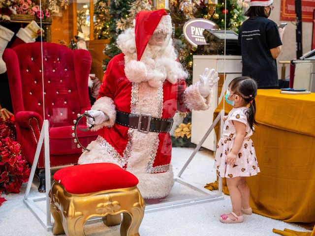 TOPSHOT - A man dressed in a Santa Claus outfit, wearing a face shield and kneeling behind a transparent barrier amid concerns over the spread of the COVID-19 coronavirus, gestures to a girl at a shopping mall with Christmas decorations in Kuala Lumpur on December 22, 2020. (Photo by Mohd RASFAN / AFP) (Photo by MOHD RASFAN/AFP via Getty Images)