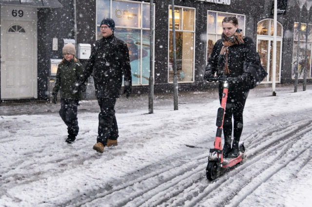 A young woman on an electric scooter drives past pedestrians on a snow-covered road during