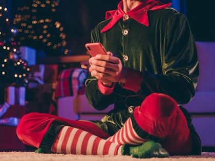 Full length photo of crazy elf sit floor hold smart phone in house, indoors with christmas x-mas noel evening illumination