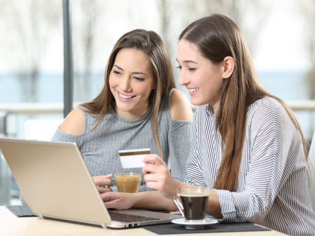 Two friends buying on line with a laptop and a credit card in a coffee shop or hotel with the sea outdoors in the background