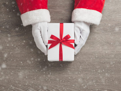 Merry Christmas concept, Top view of Santa claus hands is holding a white gift box with red ribbon and snow on wooden background.