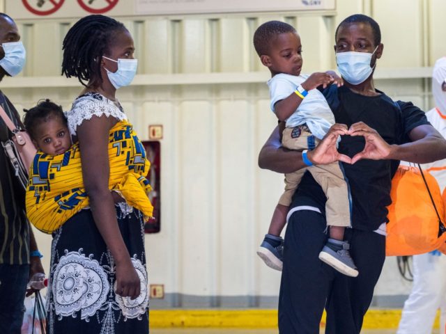 A rescued migrant carrying a baby as he stands in line forms a heart with his hands before