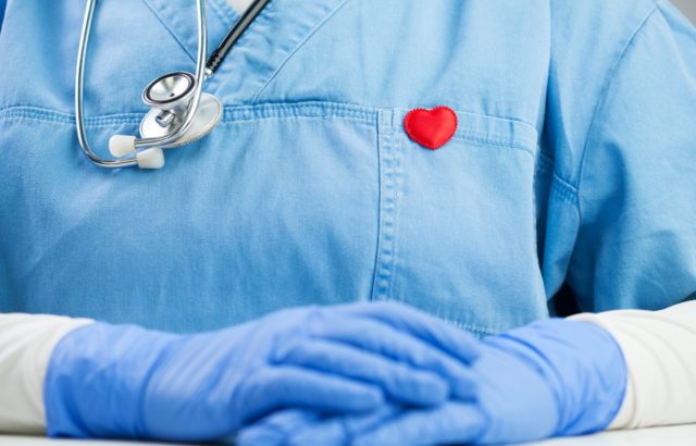 Doctor in blue uniform wearing a stethoscope and small red heart badge pin, detail closeup, GP or cardiologist in the office, sitting at the desk, Coronavirus COVID-19 global pandemic outbreak crisis