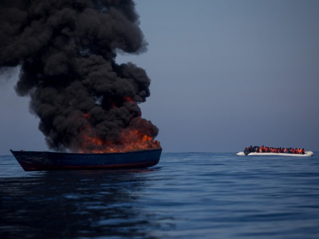 LAMPEDUSA, ITALY - MAY 18: A small rubber boat overcrowded with refugees and migrants passes a boat set alight by rescue crews from the Migrant Offshore Aid Station (MOAS) Phoenix vessel after all passengers were rescued on May 18, 2017 in the Lampedusa, Italy. Numbers of refugees and migrants attempting …