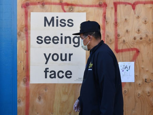 A man in a face mask walks by a sign posted on a boarded up restaurant in San Francisco, C