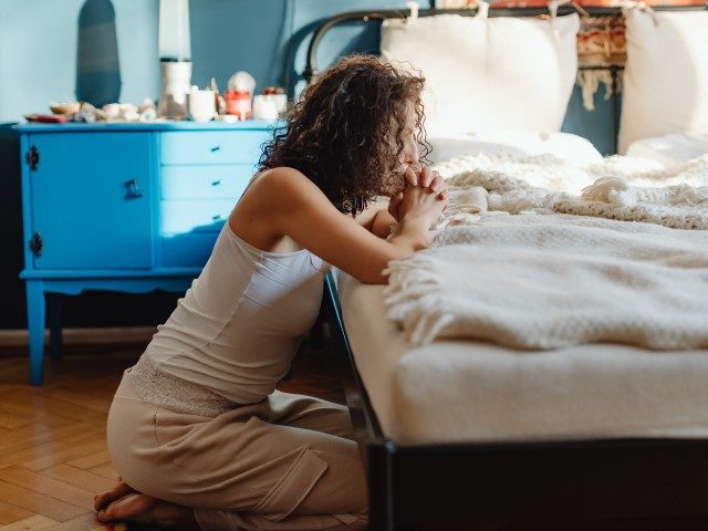 A woman prays, kneeling by a bed.