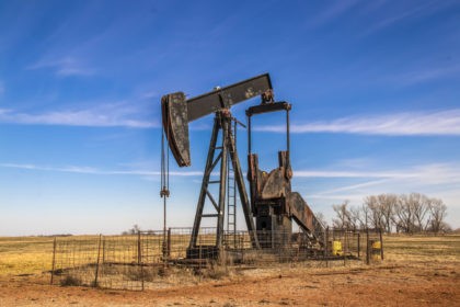 Large old rusty oil well pump jack surrounded by cattle panel fence out in field with very