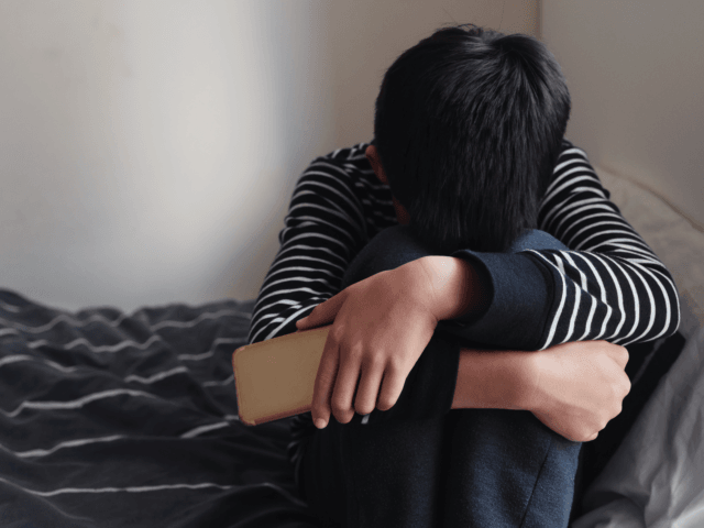 Young Asian preteen teenager boy hugging his knee in his bedroom with smartphone, Cyber bu