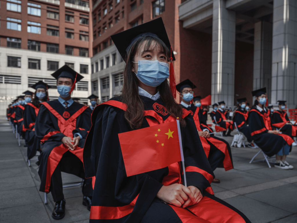 BEIJING, CHINA - JUNE 30: A Chinese student from Renmin University of China holds a flag as she sits with others as they are seated to adhere to social distancing during their graduation ceremony at the school's campus on June 30, 2020 in Beijing, China. Renmin University, also known as The Peoples University of China, is considered one of the countrys premier institutions and is one of the few campuses in Beijing this year to hold a ceremony for some of its graduating students due to concerns amid the coronavirus pandemic. The commencement, which also included virtual elements, was smaller than in previous years and students and faculty were required to wear protective masks and adhere to social distancing measures. More than 8 million students will graduate from Chinese universities and colleges this year, according to government figures. (Photo by Kevin Frayer/Getty Images)