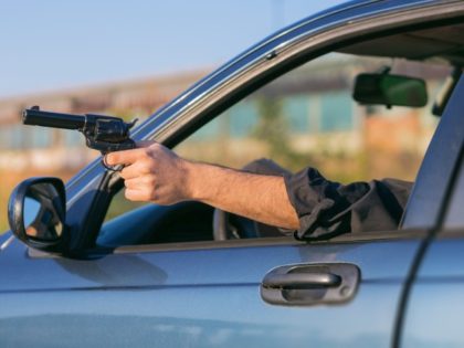 Criminal aims a gun through a window of a moving car in drive-by shooting.