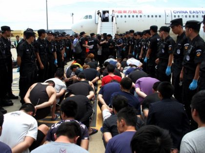 Suspects sit as Chinese police officials stand guard before boarding a plane at the Phnom