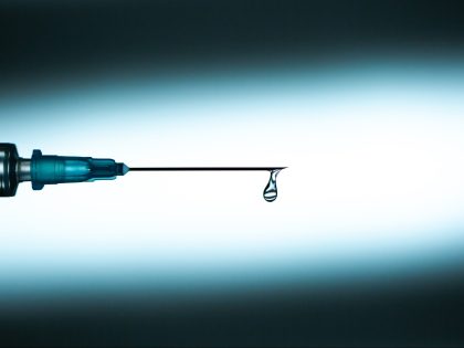 Closeup drop of clean transparent medication spilling from end of needle of disposable syringe against blue background