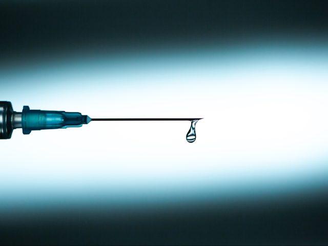 Closeup drop of clean transparent medication spilling from end of needle of disposable syr