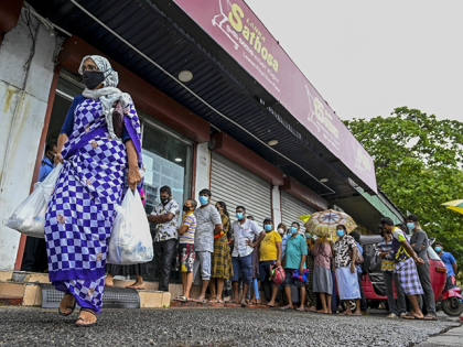A woman carrying food bags walks pasts people standing in queue outside a state-run supermarket to buy essential food items in Colombo on September 3, 2021 as Sri Lanka began imposing price controls on essential food from September 3 after using a state of emergency to seize allegedly hoarded stocks …