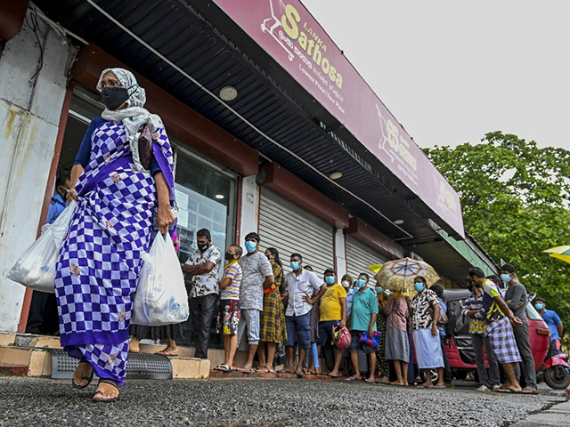 A woman carrying food bags walks pasts people standing in queue outside a state-run superm