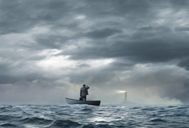 A beacon from a lighthouse beckons a stranded businessman as he stands in a small boat that floats under an ominous sky and choppy waters.