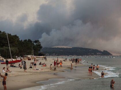 TOPSHOT - This photograph taken on July 18, 2022 shows people swim on the Moulleau's beach as the smoke rising from the forest fire in La Teste-de-Buch, seen from Arcachon, in front of the Pilat dune. - In scorching heat, with more than 40°C, some 8,000 people had to leave …