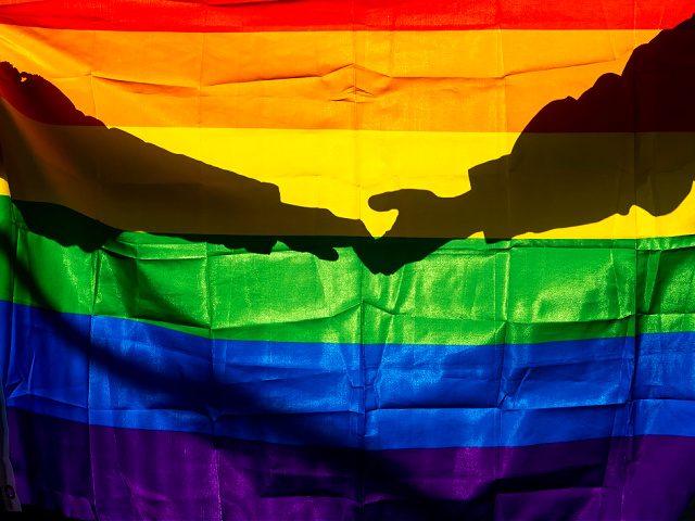 Two women are holding hands against the light through the LGTBI flag. - stock photo