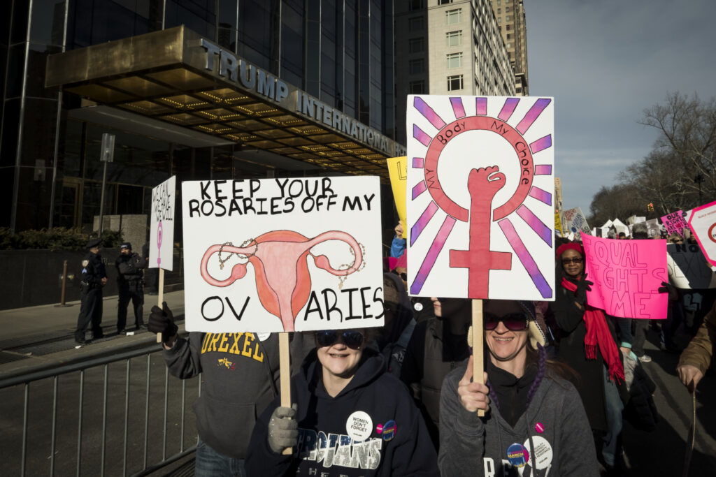 NEW YORK, NY - JANUARY 20: A demonstrator holds up a banner saying "Keep Your Rosaries Off My Ovaries" while another has a banner saying "My Body, My Choice" in front of Trump International Hotel and Tower during the second annual Women's March in the borough of Manhattan in New York City, U.S. on Saturday, January 20, 2018. One year after the inauguration of President Donald Trump, thousands of people will again gather to protest for equal rights at the 2018 Women's March. (Photo by Ira L. Black/Corbis via Getty Images)