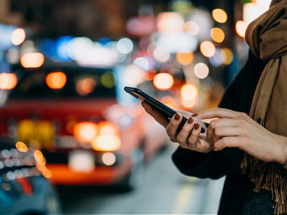 Close up of young woman using mobile app device on smartphone to arrange taxi ride in downtown city street, with illuminated busy city traffic scene during rush hour with traffic congestion in the evening - stock photo
