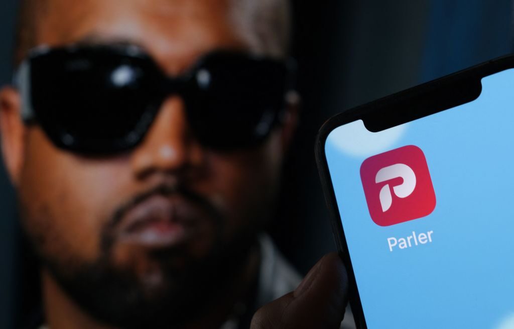 This illustration photo shows the Parler social network app logo on a cell phone screen with a picture of US rapper Kanye West in the background in Los Angeles, October 17, 2022. - Social network Parler announced on October 17 a deal for Kanye West to buy the platform popular with US conservatives, just over a week after the rapper's Twitter and Instagram accounts were restricted over anti-Semitic posts. West -- now known as Ye -- has recently alienated fans and business partners with anti-Semitic comments, interest in racist conspiracy theories and wearing a provocative "White Lives Matter" T-shirt at Paris fashion week. (Photo by Chris DELMAS / AFP) (Photo by CHRIS DELMAS/AFP via Getty Images)