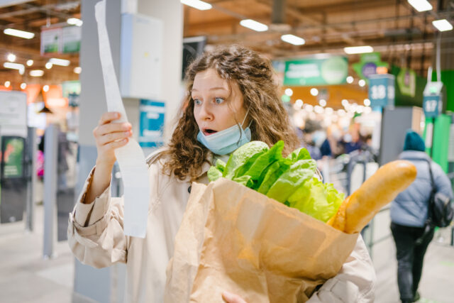 Young woman in a medical mask looks shocked at a paper check in a grocery supermarket hold