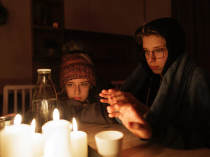 During the energy crisis family is sitting by the table, lit by candles. Everyone is wearing warm clothes because of heating problems during the power outage. They are eating dry biscuits and warming hands from the candle flame. Canon R5