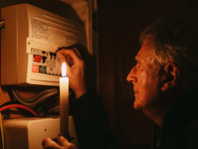 A senior man investigates his fuse box at home - by the light of a candle only - in a blac