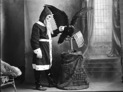 A man wearing a Santa Claus suit and mask, holding his finger out to a stuffed bird of prey, which is holding an American flag, Gray, Oklahoma, circa 1910. (Photo by Ward Brothers Studio/Oklahoma Historical Society/Getty Images)